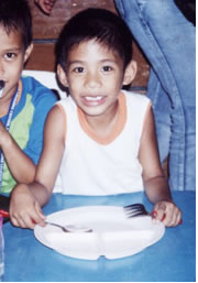 Happy boy holding spoon and fork, smiling expectantly before empty plate.